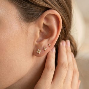 Könnte beinhalten: Drei goldene Blumen-Ohrstecker mit kleinen, funkelnden Steinen werden an einem Ohr gezeigt. Die Ohrringe sind unterschiedlich groß, der größte unten und der kleinste oben. Das Ohr ist vor einem verschwommenen Hintergrund.