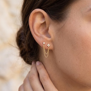 May include: A gold double-chain earring featuring a single round stud and a four-stone flower-shaped stud connected by two delicate gold chains that drape beneath the earlobe, creating a layered look with clear sparkling stones.