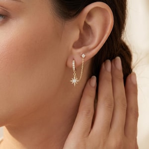 Könnte beinhalten: Goldfarbene Ohrringe mit einem sternförmigen Anhänger und einem kleinen Stecker, verbunden durch eine feine Kette. Der Creolen-Ohrring ist mit kleinen, funkelnden Steinen besetzt. Der Sternanhänger und der Stecker haben ebenfalls ähnliche Steine.
