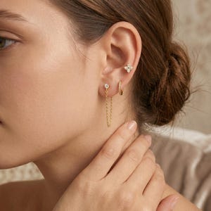 May include: A close-up of an ear displaying several gold earrings. The selection includes a small hoop, a stud with a chain, and a four-petal flower design. Each earring is decorated with tiny, sparkling gemstones.