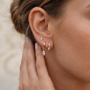 May include: A set of three gold-toned earrings worn in one ear. The top earring is a small marquise-cut stud. The middle earring is a gold hoop with a dangling marquise-cut stone. The bottom earring is a small hoop featuring three small stones.