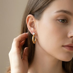 May include: Gold-colored hoop earrings with a clear, round gemstone at the top. The earrings are a polished gold and have a classic, minimalist design. The model is wearing the earrings in her ear.