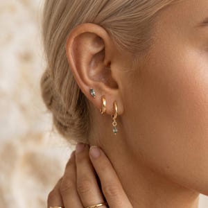 May include: A set of gold-toned earrings consisting of a small stud with a marquise-cut blue-green gemstone, a plain gold hoop, and a gold hoop featuring a dangling marquise-cut blue-green gemstone. The jewelry is worn in three ear piercings.