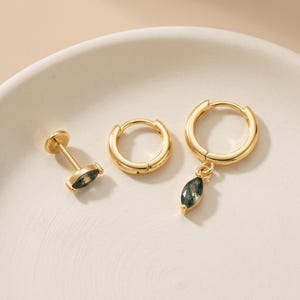 Puede incluir: Un conjunto de tres piezas de joyería en tono dorado que incluye un pendiente de botón con parte posterior plana y una piedra de ágata musgosa verde en talla marquesa, un aro pequeño liso y un aro con un dije de ágata musgosa verde.