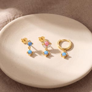 May include: Three gold-tone earrings featuring opal-like stones. One stud has two blue round stones, one stud features a pink round stone with white accents and a blue drop, and one hoop earring has a single blue round stone dangling from the base.