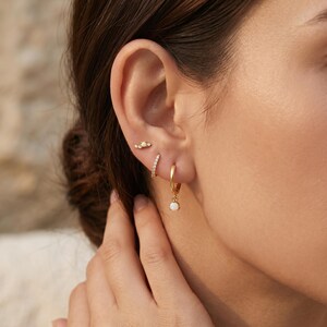 May include: Three gold earrings worn in one ear. The top earring is a small curved bar with three tiny white stones. The middle earring is a thin gold hoop encrusted with small clear stones. The bottom earring is a gold hoop with a single white stone.