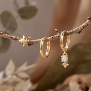 May include: A set of three gold-toned earrings hanging from a branch. One earring is a four-pointed star stud, one is a plain circular hoop, and the third is a circular hoop featuring a dangling white opal stone topped with a four-pointed star charm.