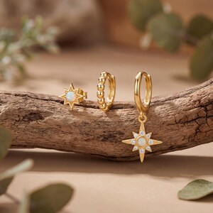 May include: A set of gold-toned jewelry consisting of a star-shaped stud earring with a central opal, a small beaded hoop earring, and a larger hoop earring featuring a dangling star-shaped charm with a central opal and small clear stones.