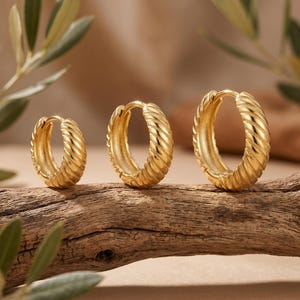 May include: Three gold-colored hoop earrings with a ridged design are displayed on a piece of wood. The earrings vary in size, with the largest on the right and the smallest on the left. The background includes green leaves and a blurred neutral backdrop.
