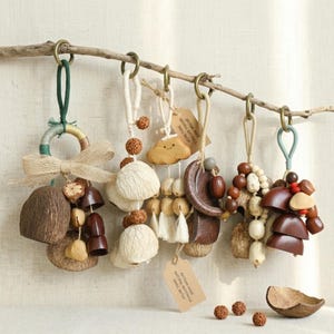 May include: A collection of handmade natural material ornaments hanging from a branch. The ornaments feature various shapes and textures, including wood, beads, and tassels. Colors include brown, beige, and cream. Each ornament is unique, with a rustic and organic aesthetic.