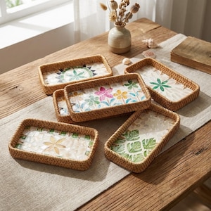 Könnte beinhalten: Eine Sammlung rechteckiger Tabletts aus geflochtenem Rattan mit dekorativen Mosaikeinsätzen. Die Tabletts zeigen verschiedene Blumen- und Blättermuster in Grün-, Rosa- und Gelbtönen. Die Tabletts sind auf einem Holztisch mit einem neutralfarbenen Tischläufer angeordnet.