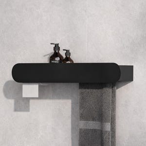 May include: A matte black wall-mounted bathroom shelf with rounded ends. The shelf holds two amber glass pump bottles labeled Aesop. A white toilet paper roll hangs from the left side, and a folded gray towel hangs from the right side.