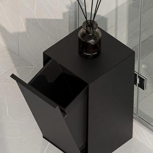 May include: A matte black rectangular trash bin with a tilt-out front panel and a flat top surface. A dark glass reed diffuser bottle with black reeds and the text ETERNAL MUSK printed on the front sits on top of the bin.