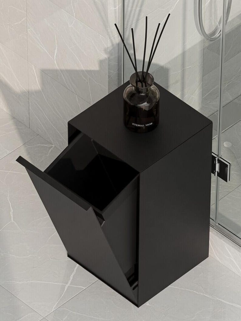May include: A matte black rectangular trash bin with a tilt-out front panel and a flat top surface. A dark glass reed diffuser bottle with black reeds and the text ETERNAL MUSK printed on the front sits on top of the bin.