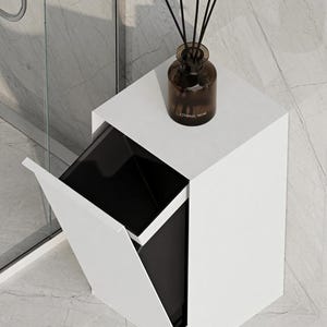 May include: A white rectangular trash bin with a tilting front panel that reveals a black inner container. A brown glass bottle with black reed diffuser sticks sits on the flat top surface of the bin. The bin has a clean, solid white exterior.