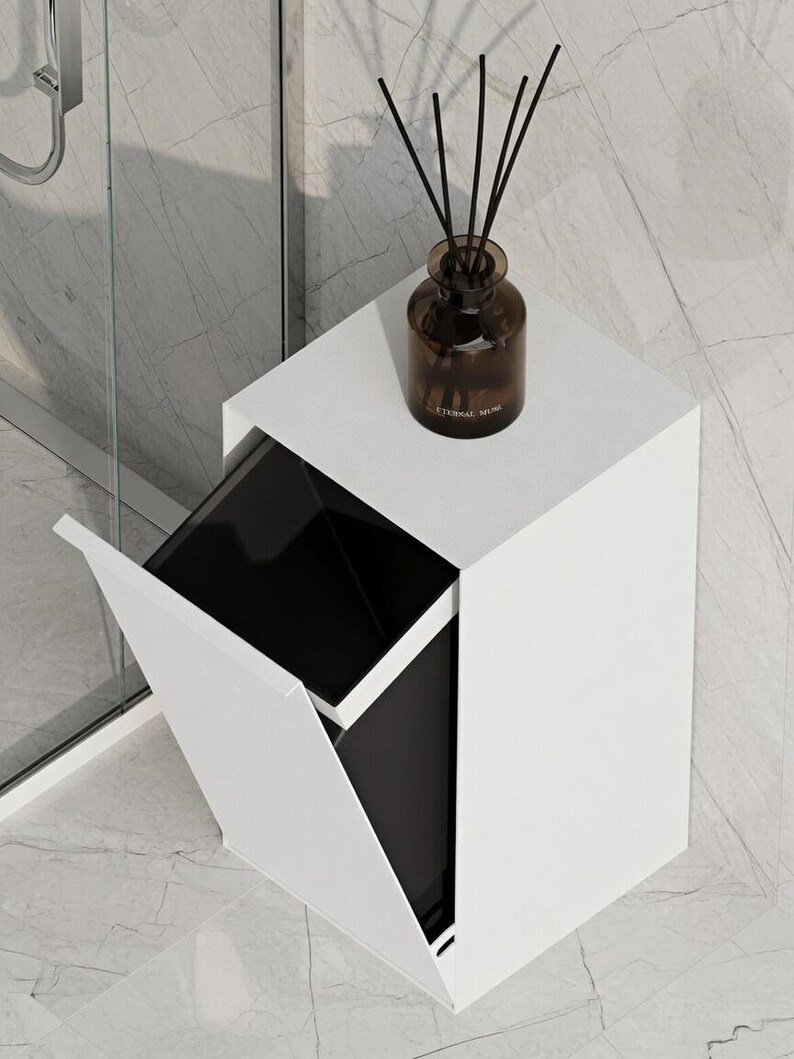 May include: A white rectangular trash bin with a tilting front panel that reveals a black inner container. A brown glass bottle with black reed diffuser sticks sits on the flat top surface of the bin. The bin has a clean, solid white exterior.
