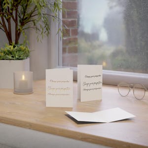 Könnte beinhalten: Zwei weiße, gefaltete Grußkarten mit dem in Gold- und Schwarztinte gedruckten Text Change your perspective. Eine Karte steht aufrecht und zeigt den Text, während eine andere flach liegt. Eine Brille mit dünnem Rahmen liegt auf der Holzoberfläche.