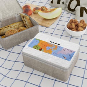Könnte beinhalten: Eine rechteckige Lunchbox mit einer strukturierten hellgrauen Außenseite und einem weißen Deckel, der eine farbenfrohe Karte von Nordamerika und den in Schwarz gedruckten Namen Noah zeigt. Die Lunchbox steht auf einer weißen Fläche mit blauem Gittermuster.