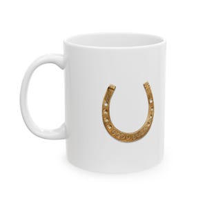Könnte beinhalten: Ein weißer Keramikbecher mit einer mittigen Grafik eines metallisch goldenen Hufeisens. Das Hufeisen ist mit der Öffnung nach oben ausgerichtet und enthält den geprägten Text GOOD LUCK entlang der unteren Kurve der Metalloberfläche.