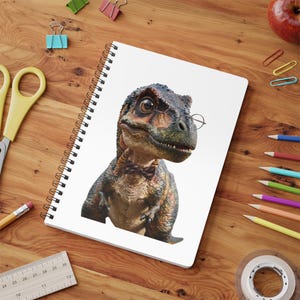 Peut inclure: Un carnet à spirales présentant l'illustration d'un dinosaure brun et vert portant des lunettes rondes à monture métallique et un petit nœud papillon rayé. Le carnet est blanc avec une reliure à spirale noire sur une surface en bois.