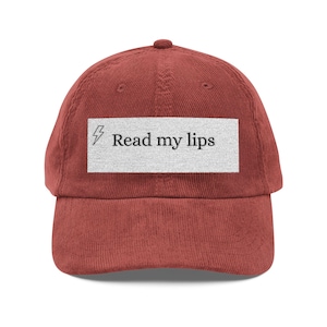 Könnte beinhalten: Eine rostfarbene Baseballkappe aus Cord mit einem rechteckigen weißen Aufnäher auf der Vorderseite. Der Aufnäher zeigt ein kleines schwarzes Blitzsymbol, gefolgt von dem schwarzen Text "Read my lips" in einer klaren Serifenschrift.