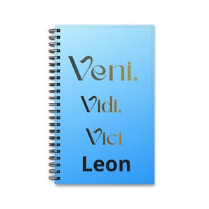 Könnte beinhalten: Ein spiralgebundenes Notizbuch mit einem hellblauen Farbverlauf auf dem Einband. Auf der Vorderseite steht der Text Veni, Vidi, Vici in einer dunklen, stilisierten Schrift, gefolgt von dem Namen Leon in einer fetten, schwarzen serifenlosen Schrift.