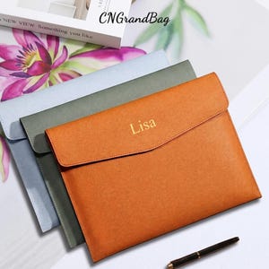 May include: Three rectangular document sleeves with a textured finish, stacked in orange, olive green, and light blue. The orange sleeve features the name Lisa embossed in gold lettering on the front flap. A black pen lies nearby.