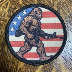 May include: A circular patch depicting a muscular Bigfoot figure with a rifle, set against a backdrop of the American flag. The design incorporates blue, white, and red, with a black border.