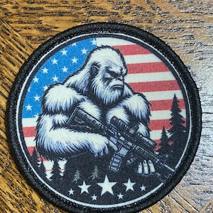 May include: Round patch featuring a white Bigfoot-like creature holding a rifle, set against a backdrop of the American flag. The design includes stars, stripes, and silhouettes of trees, all framed by a black border.
