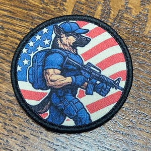 Könnte beinhalten: Runder Aufnäher mit einem Schäferhund in blauer Uniform und Mütze, der ein Gewehr hält. Der Hintergrund hat ein amerikanisches Flaggen-Design mit roten und weißen Streifen und einem blauen Sternenfeld. Schwarzer Rand.