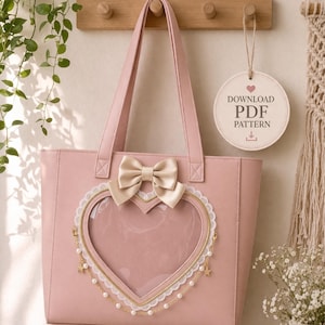May include: A pink tote bag with a heart-shaped window, adorned with a cream-colored bow and lace trim. The bag features a gold-colored frame around the heart and hanging gold charms. A tag reads "DOWNLOAD PDF PATTERN".