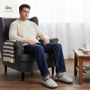 May include: A person seated in a gray armchair wearing a cream-colored sweater and blue jeans. They are wearing gray and white striped slippers. A striped blanket is draped over the chair. A small wooden table with a mug is to the right.
