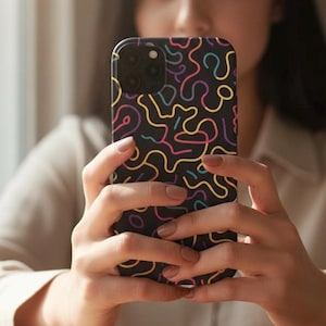 Può includere: Custodia nera per smartphone con un colorato motivo astratto di linee ondulate in giallo, rosa, blu e viola. La custodia ha una finitura opaca e copre la parte posteriore e i lati del telefono. La custodia del telefono è tenuta da una persona.