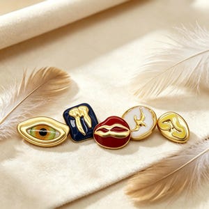 Puede incluir: Cinco botones decorativos de metal con bordes dorados y detalles en esmalte. Los diseños incluyen un ojo realista, un diente dorado sobre fondo azul oscuro, labios rojos, un óvalo blanco con un pájaro dorado y una forma abstracta amarilla.