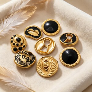 May include: A collection of eight gold-toned metal buttons with black accents. The buttons feature various designs including a keyhole, a pair of lips, a spiral, and an eye motif. Some buttons have smooth black centers while others are engraved.