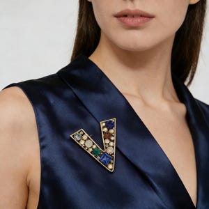 Peut inclure: Broche dorée en forme de lettre "V" ornée de pierres précieuses colorées. La broche est posée sur un vêtement en satin bleu marine. Les pierres précieuses comprennent des pierres bleues, vertes et claires.