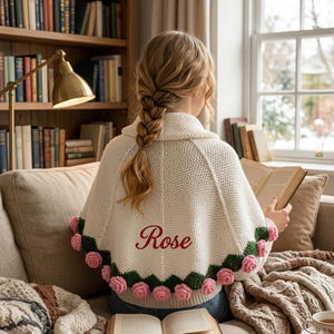 Könnte beinhalten: Eine cremefarbene gehäkelte Kapuze mit gewelltem Rand, verziert mit rosa Rosenapplikationen und grünen Blättern. Das Wort "Rose" ist rot gestickt. Die Kapuze wird von einer Person getragen, die ein Buch liest.