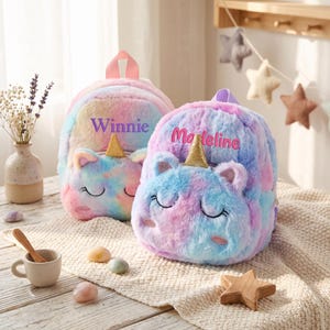 May include: Two plush unicorn backpacks with soft, multi-coloured pastel fur in shades of pink, blue, and yellow. Each bag features a gold glitter horn, embroidered closed eyes, and pink cheeks. The left bag is personalised with the name Winnie, and the right bag is personalised with the name Madeleine in pink script.