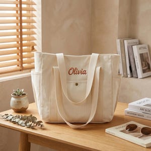 Puede incluir: Bolso tote de lona color crema con correas largas y bolsillos laterales. La palabra "Olivia" está bordada en escritura roja en la parte delantera. La bolsa está sobre una mesa de madera junto a una pequeña planta suculenta y una revista.
