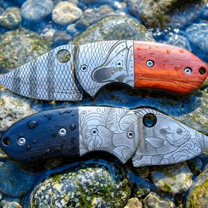 May include: Two folding pocket knives with metal blades and wooden handles. The top knife features a cross-hatched blade pattern and a reddish-brown handle. The bottom knife displays an engraved fish design on the blade and a dark black handle.