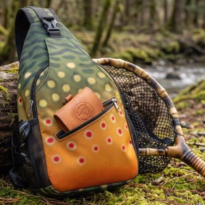 May include: A sling backpack featuring a brown trout skin pattern with green, yellow, and red spots. The bag has a black mesh side pocket, a black adjustable strap with a buckle, and a tan leather wallet with the text Graywolf Fly Fishing.