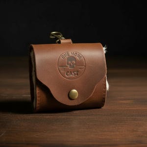 Może przedstawiać: Brązowe skórzane etui z mosiężnym zapięciem i zatrzaskiem. Etui posiada okrągły emblemat z czaszką i napisem "ONE MORE CAST". Wnętrze wykonane jest z białego, teksturowanego materiału.