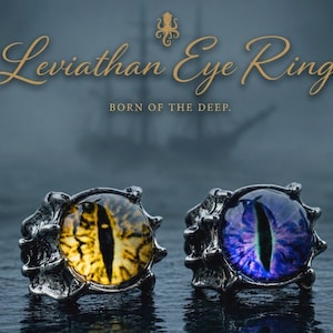 May include: Two rings with dark metallic bands featuring textured claw-like settings. One ring holds a yellow eye cabochon with a vertical black pupil, and the other holds a purple eye cabochon. Text reads: Leviathan Eye Rings BORN OF THE DEEP.