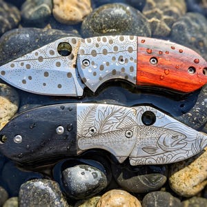 May include: Two folding pocket knives with metallic blades and handles. The top knife features a spotted pattern on the blade and a reddish-brown wood handle. The bottom knife displays an engraved fish and fern design with a black handle.
