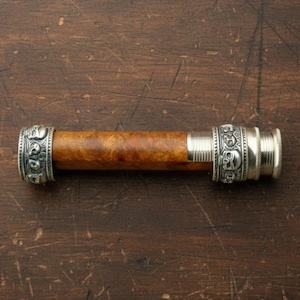 May include: A cylindrical object with a wooden body and silver-coloured metal accents. The metal bands feature skull designs. The wood has a warm, brown tone with a unique grain pattern.