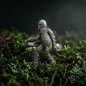 May include: A silver-toned metal ring featuring a detailed, textured figure of a hairy, upright creature resembling a Bigfoot or Sasquatch. The figure is integrated into the band, showcasing intricate fur-like carvings and a defined facial structure.