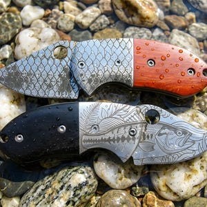 May include: Two folding pocket knives with metallic blades and handles. The top knife features a fish scale pattern on the blade and a reddish-brown wooden handle. The bottom knife displays a detailed fish illustration and a black handle.
