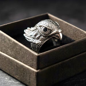 May include: A silver-toned metal ring shaped like the head of a bird of prey, featuring intricate feather detailing and a small, round black stone set as the eye. The ring is displayed inside a square, dark-colored cardboard jewelry box.