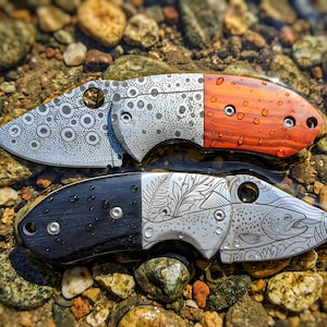 May include: Two folding pocket knives with metallic blades and wooden handles. The top knife features a circular pattern on the blade and a reddish-brown wood handle. The bottom knife displays a fish and fern engraving with a black wood handle.