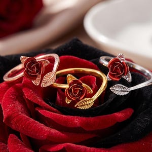 May include: Three open-style rings featuring a deep red rose blossom and a textured leaf design. The rings are finished in three distinct metallic tones: rose gold, yellow gold, and silver. Each ring has a flexible band that allows for adjustment.
